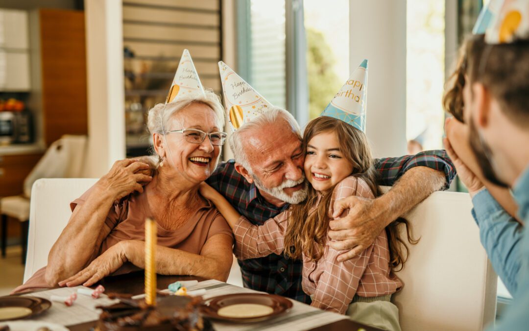 Shot of an extended family surprising their small girl with a birthday cake at home. A multigeneration family wears birthday caps, sings a song, and hugs a girl while celebrating a birthday.